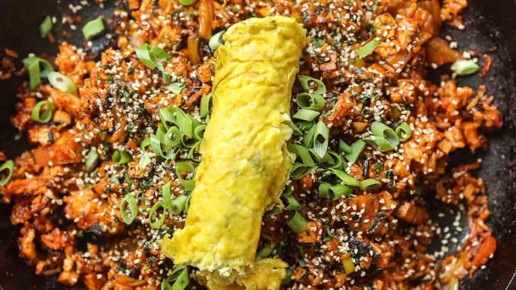 A close-up of vegan kimchi fried rice topped with a rolled omelette. The dish is garnished with sliced green onions and sesame seeds. The rice appears spicy, with a rich red hue, and is served on a dark plate.